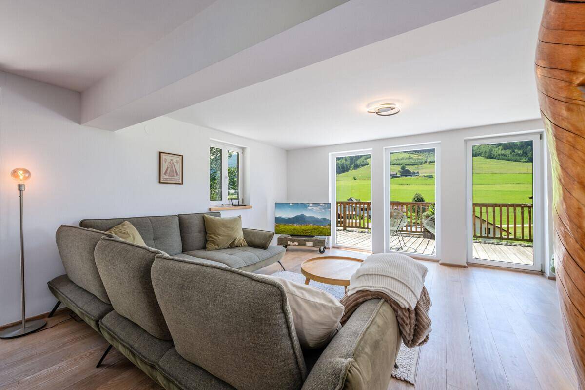 Helles Wohnzimmer mit großem Sofa, Fernseher und Zugang zum Balkon mit grüner Aussicht.