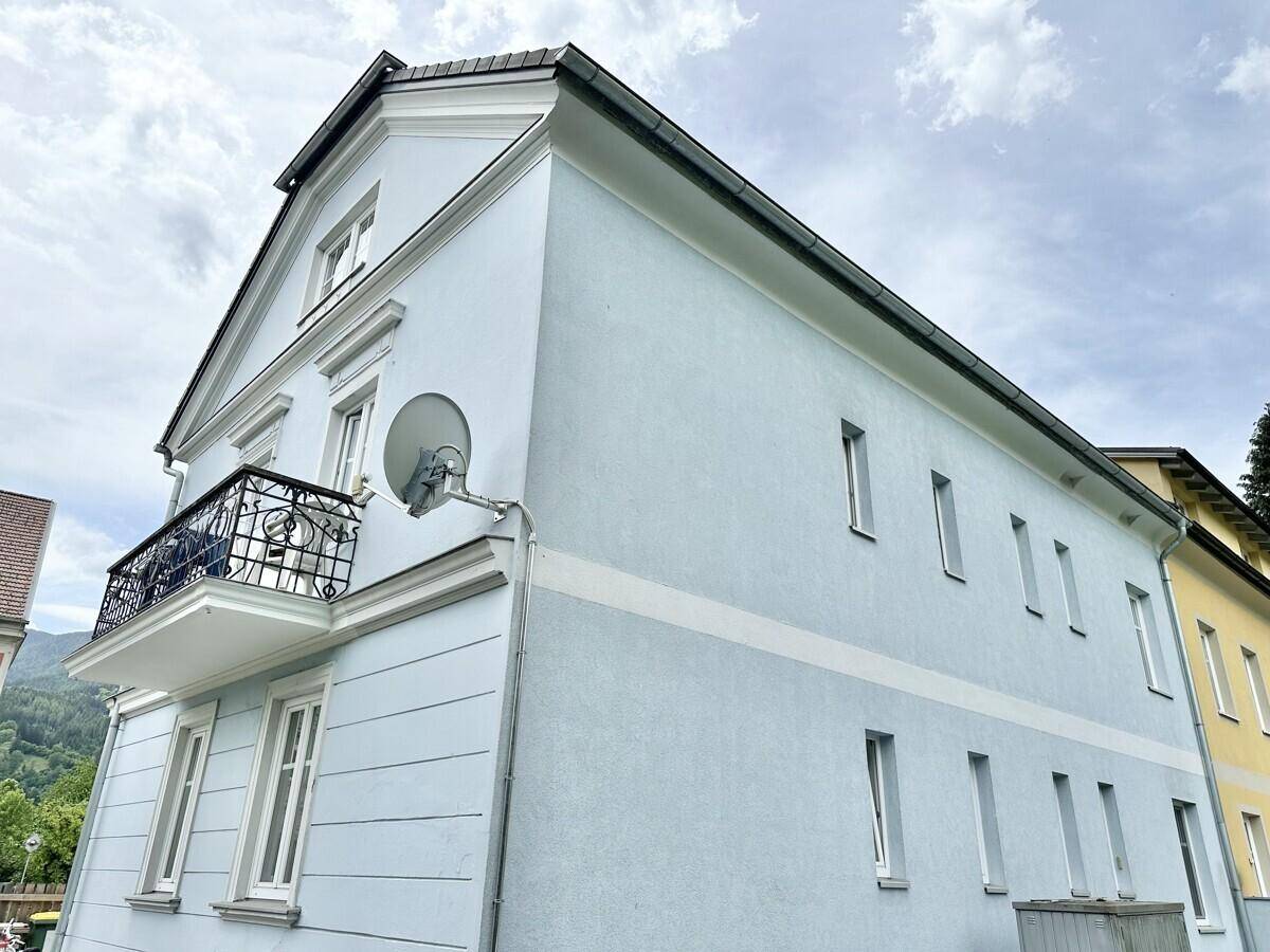 Seitenansicht des hellblauen Gebäudes mit Balkon und Satellitenschüssel, umgeben von Natur.