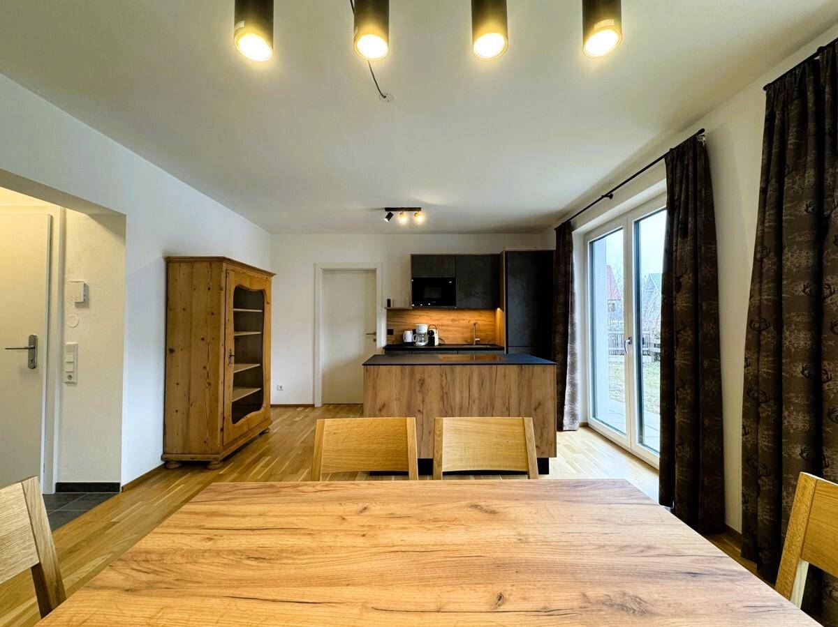 Essbereich mit Holztisch und moderner Beleuchtung, offener Zugang zur Küche und großen Fenstern.