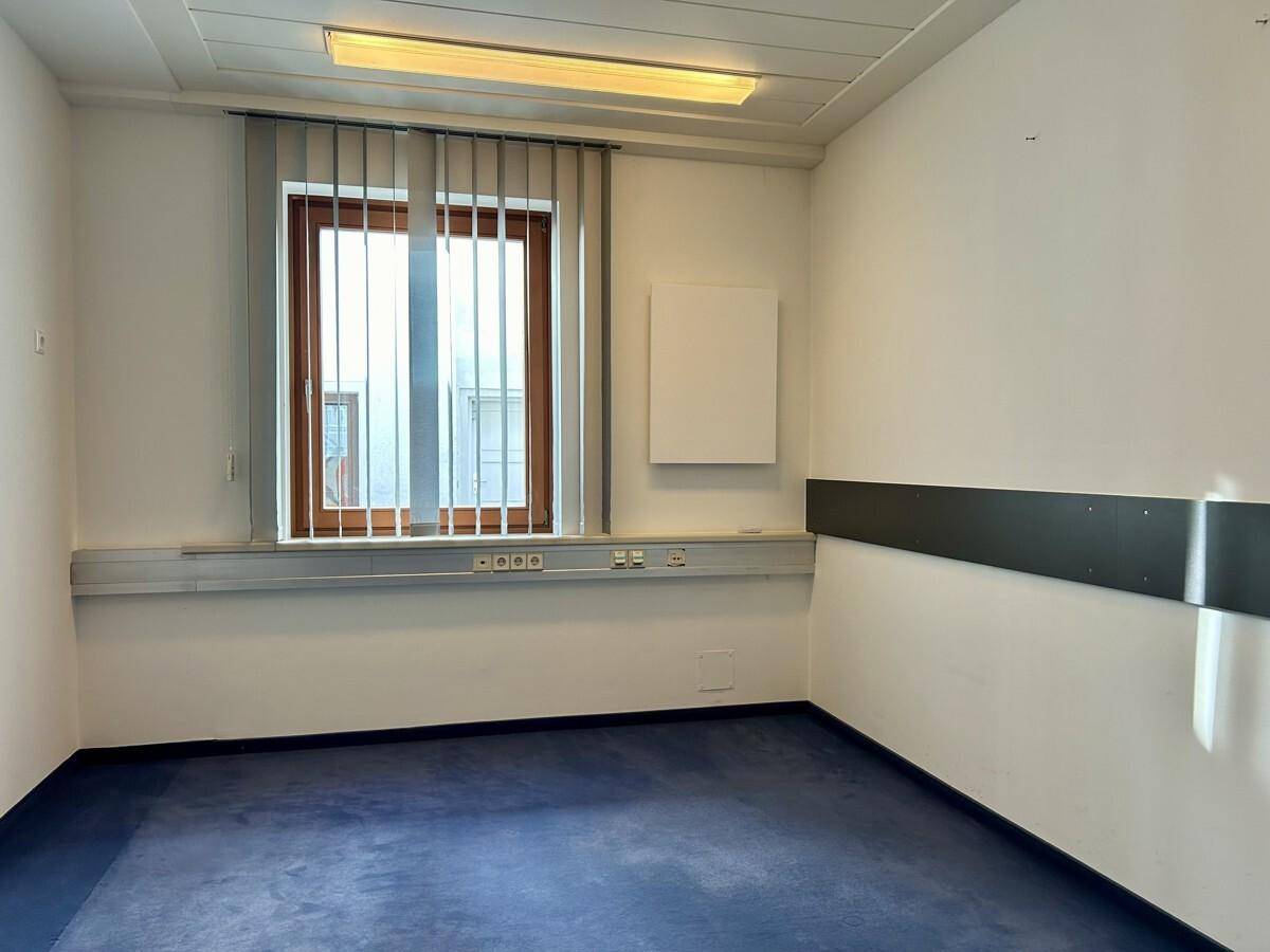 Schlichter Büroraum mit blauem Teppichboden, einem Fenster mit Jalousien und weißen Wänden.