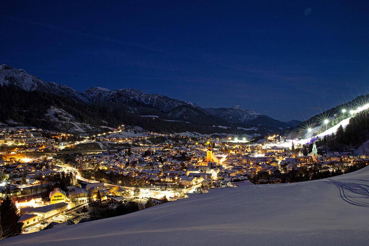 Panoramablick auf eine verschneite Bergstadt bei Nacht mit beleuchteten Skipisten und Häusern.