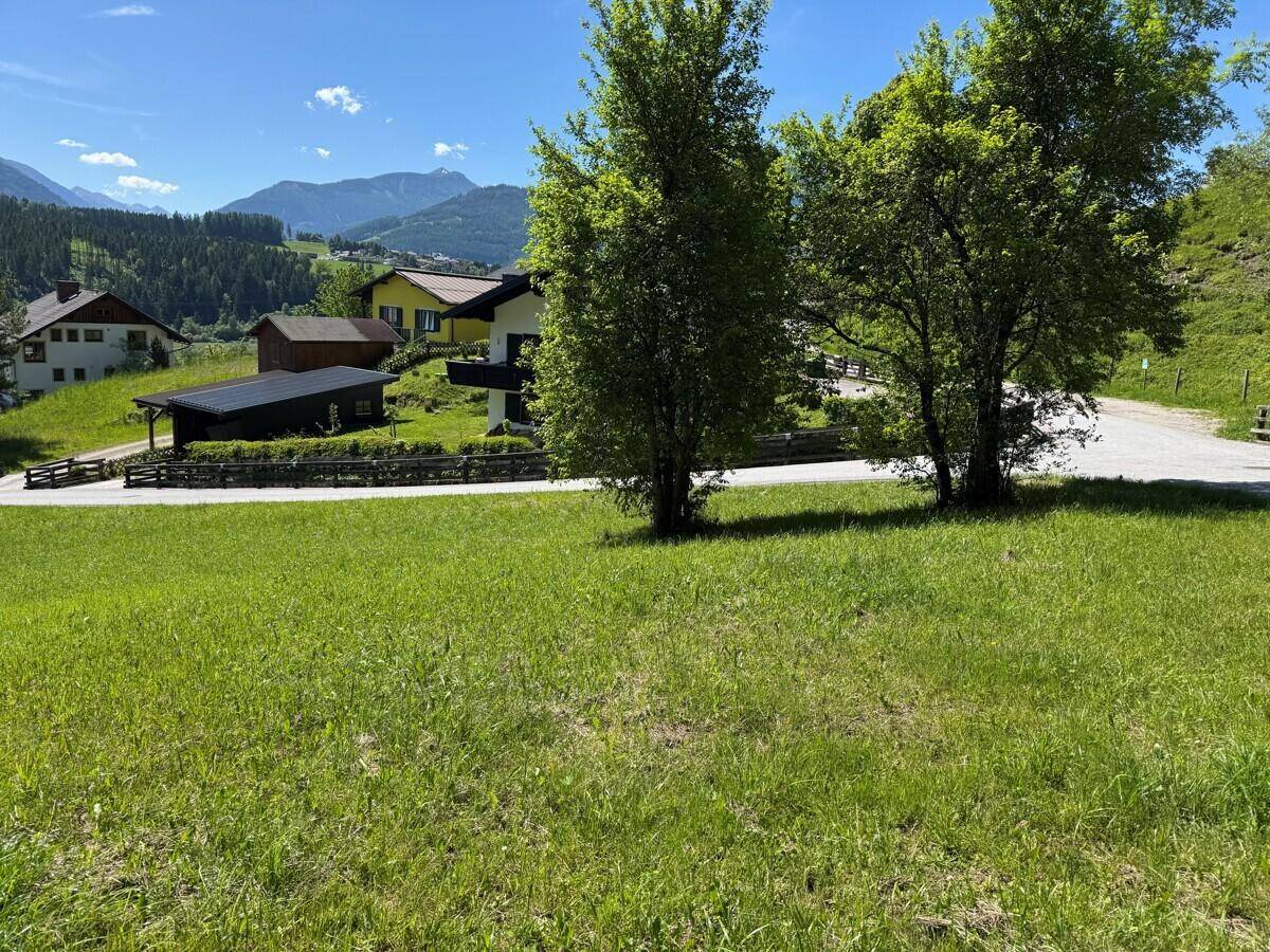 Grünes Grundstück mit Bäumen, umgeben von Häusern und einer malerischen Berglandschaft.