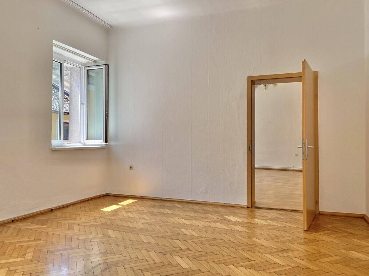 Leeres Zimmer mit Holzboden und einem geöffneten Fenster, das viel Tageslicht hereinlässt.