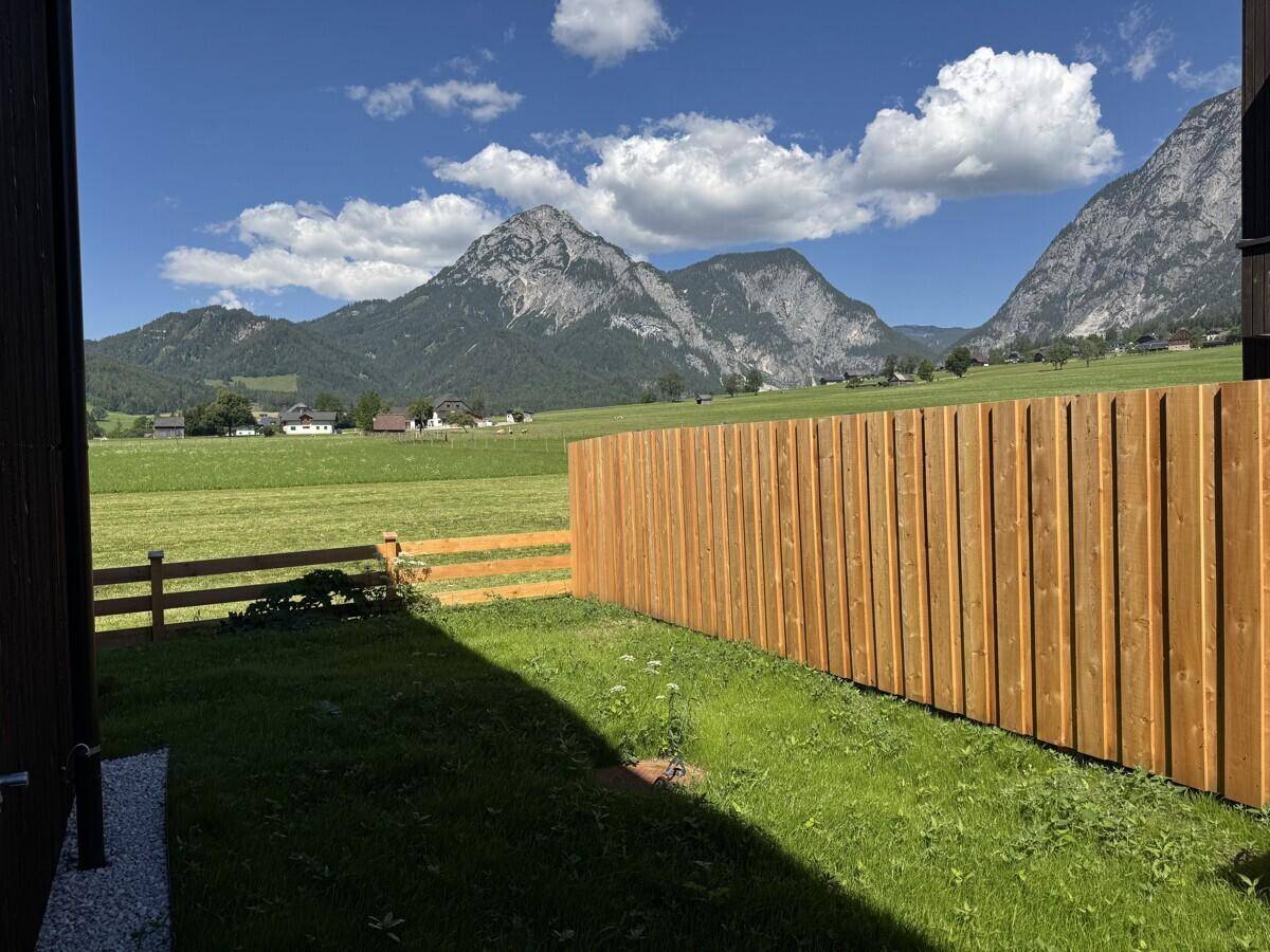 Grüner Gartenbereich mit einem hohen Holzzaun und einem beeindruckenden Bergpanorama im Hintergrund.