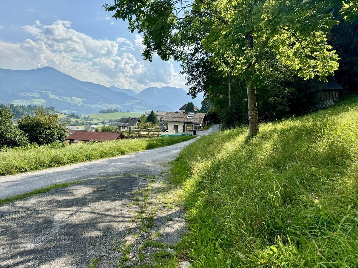 Landstraße führt bergauf mit Häusern und einer malerischen Bergkulisse im Hintergrund.