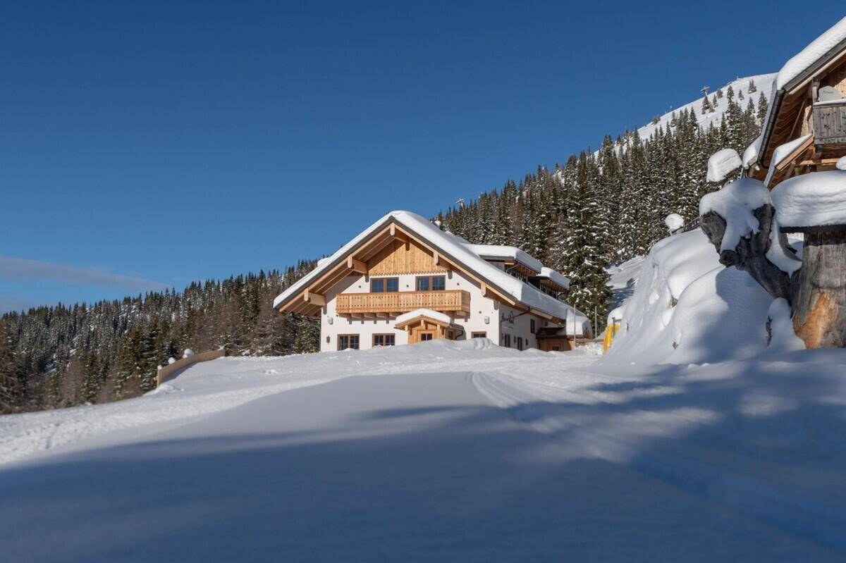 Alpenhaus mit schneebedecktem Dach und Balkonen, umgeben von verschneiten Bäumen und Hängen.