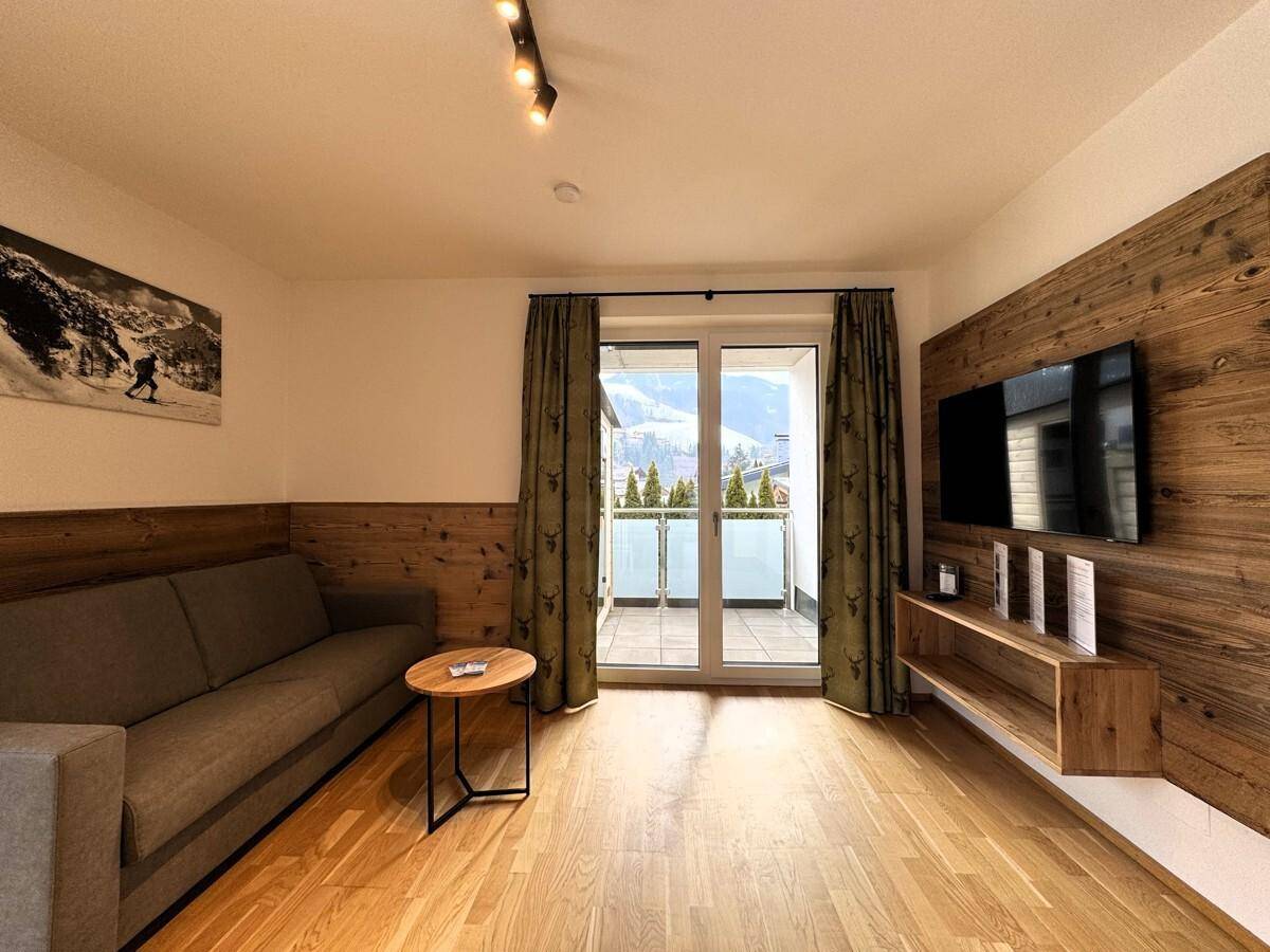 Gemütliches Wohnzimmer mit Sofa, Holzwand und Zugang zum Balkon mit Bergblick.