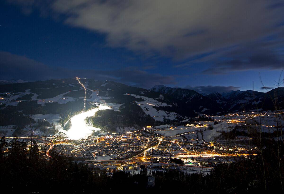 Weitläufiger Nachtblick auf eine beleuchtete Stadt im Tal, umgeben von schneebedeckten Bergen und Skipisten.