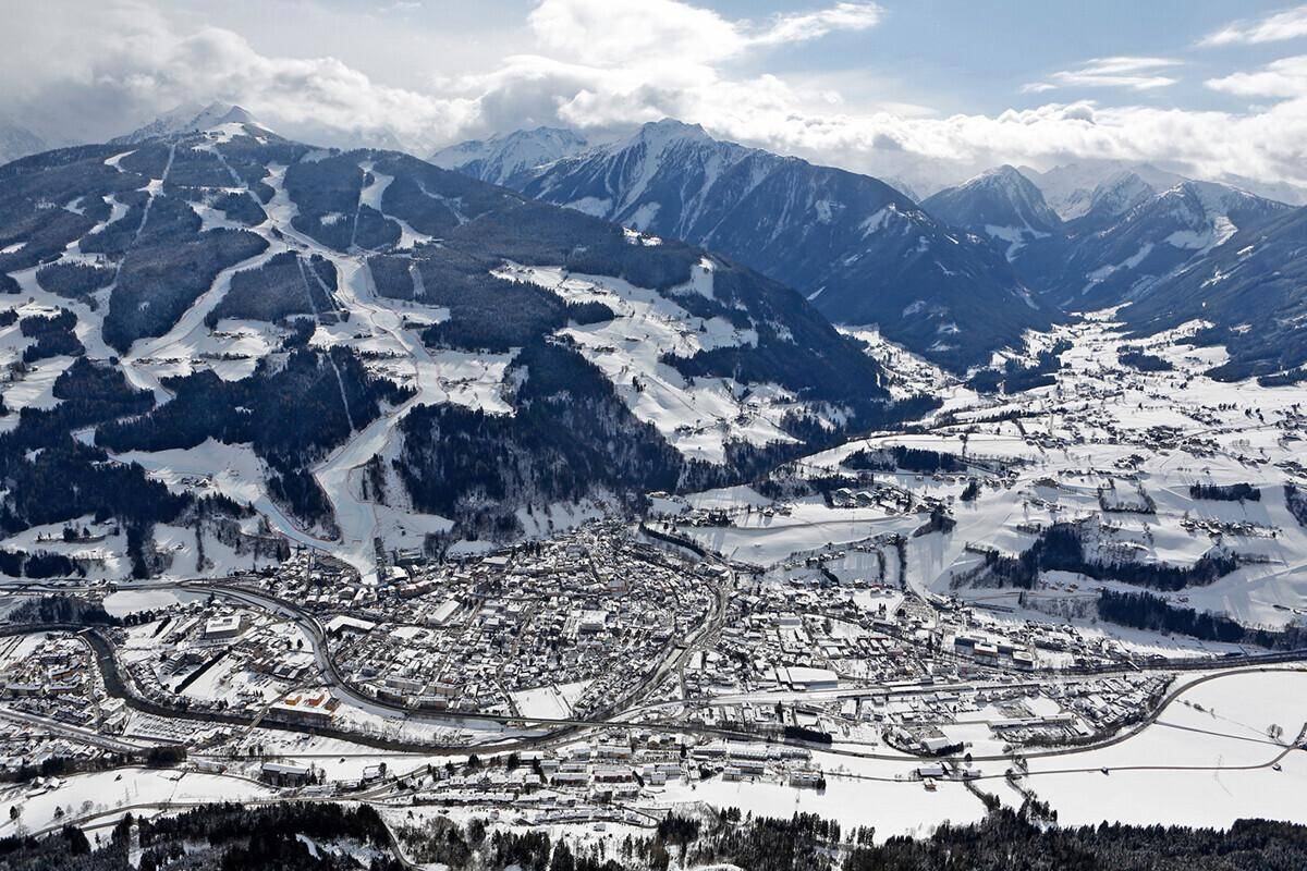 Luftaufnahme einer schneebedeckten Stadt in einem Tal, umgeben von weiten Skigebieten und majestätischen Bergen.