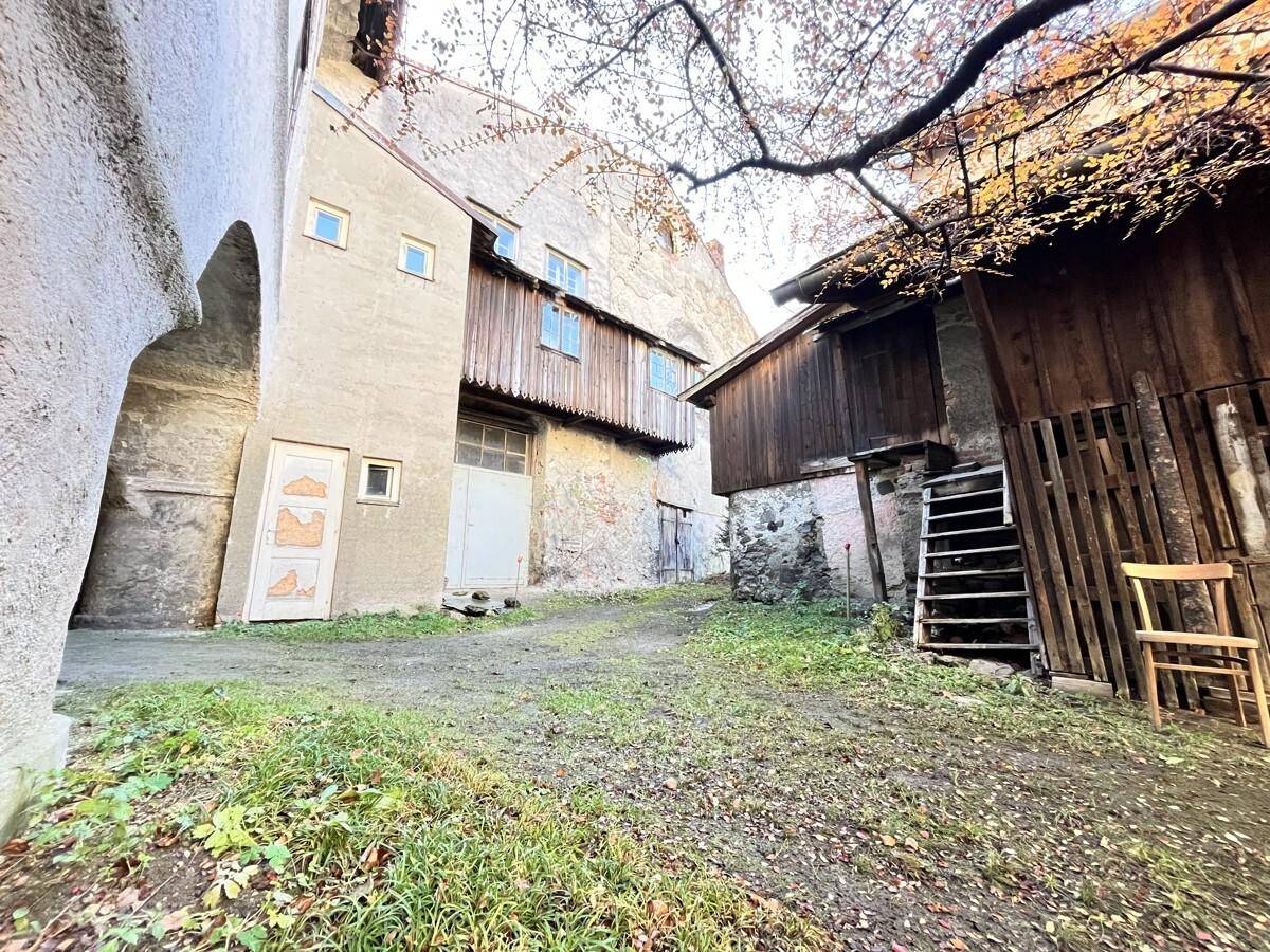 Innenhof zwischen zwei alten Gebäuden mit Holzanbauten und einem unebenen, grasbewachsenen Boden.