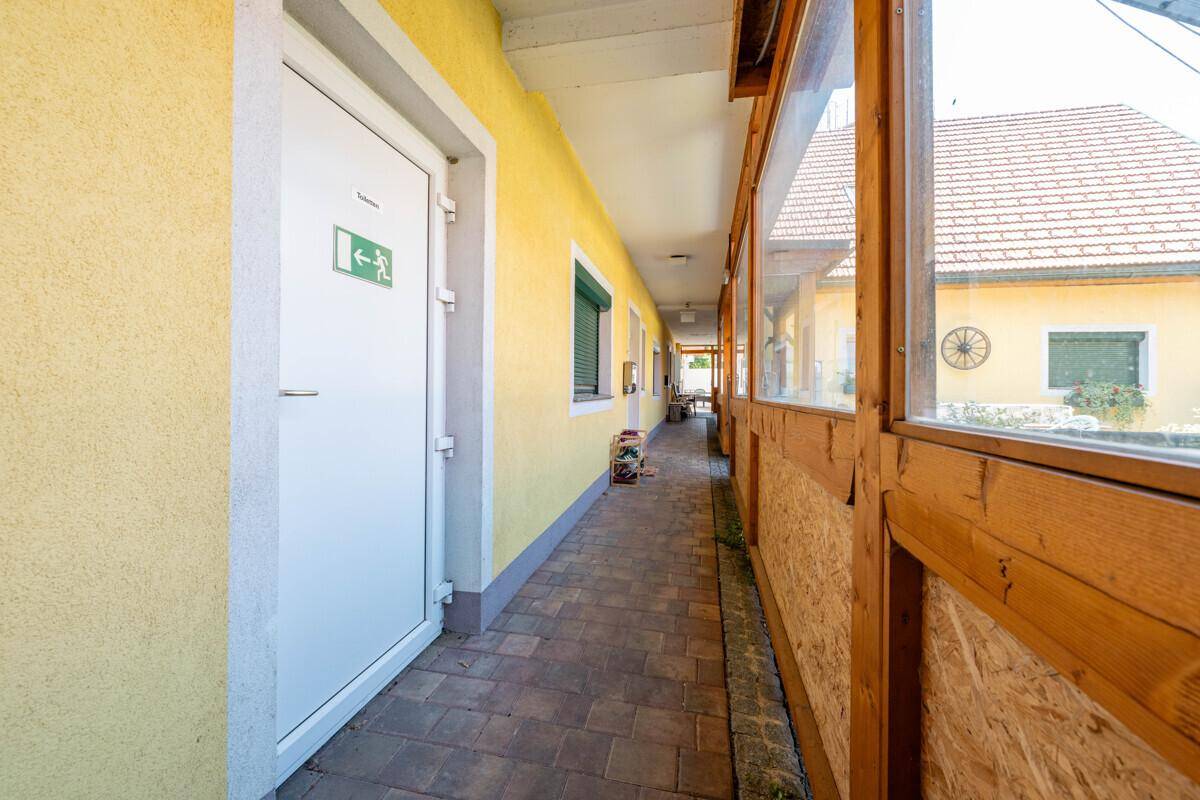 Langer, überdachter Gang mit gelber Wand und Holzelementen, führt zu weiteren Räumen.