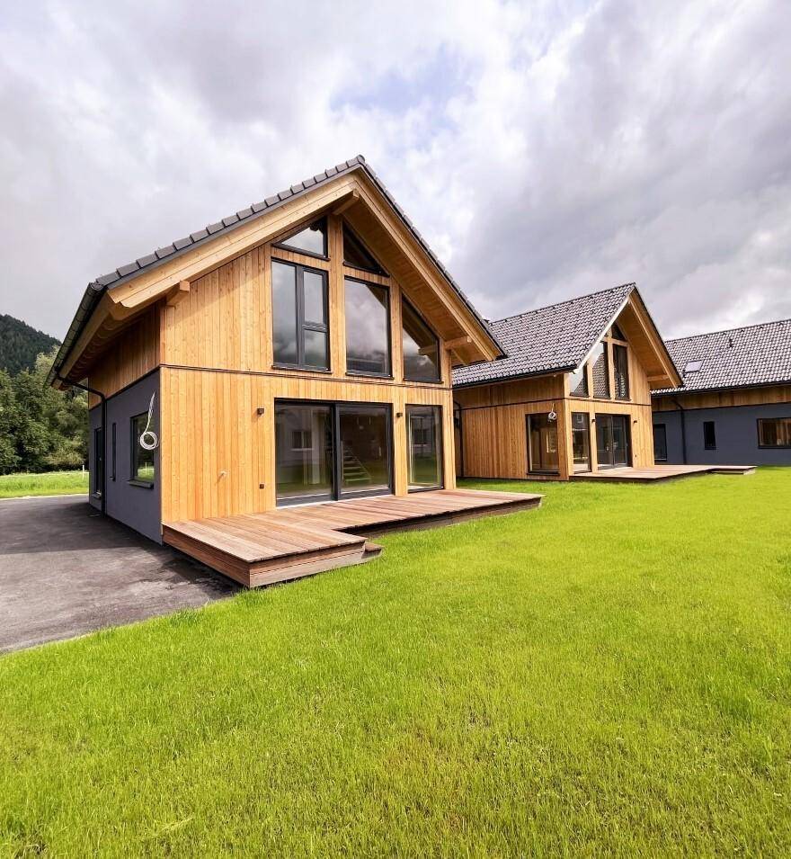Zwei moderne Einfamilienhäuser mit Holzterrassen, großen Glasfronten und weitläufigem Rasen.