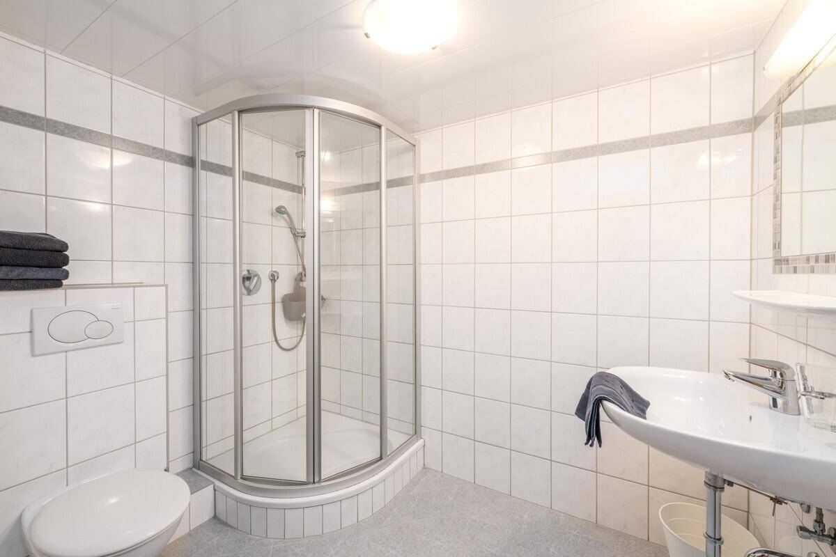 Badezimmer mit Eckdusche, weißen Fliesen und einem einzelnen Waschbecken, sauber und hell.