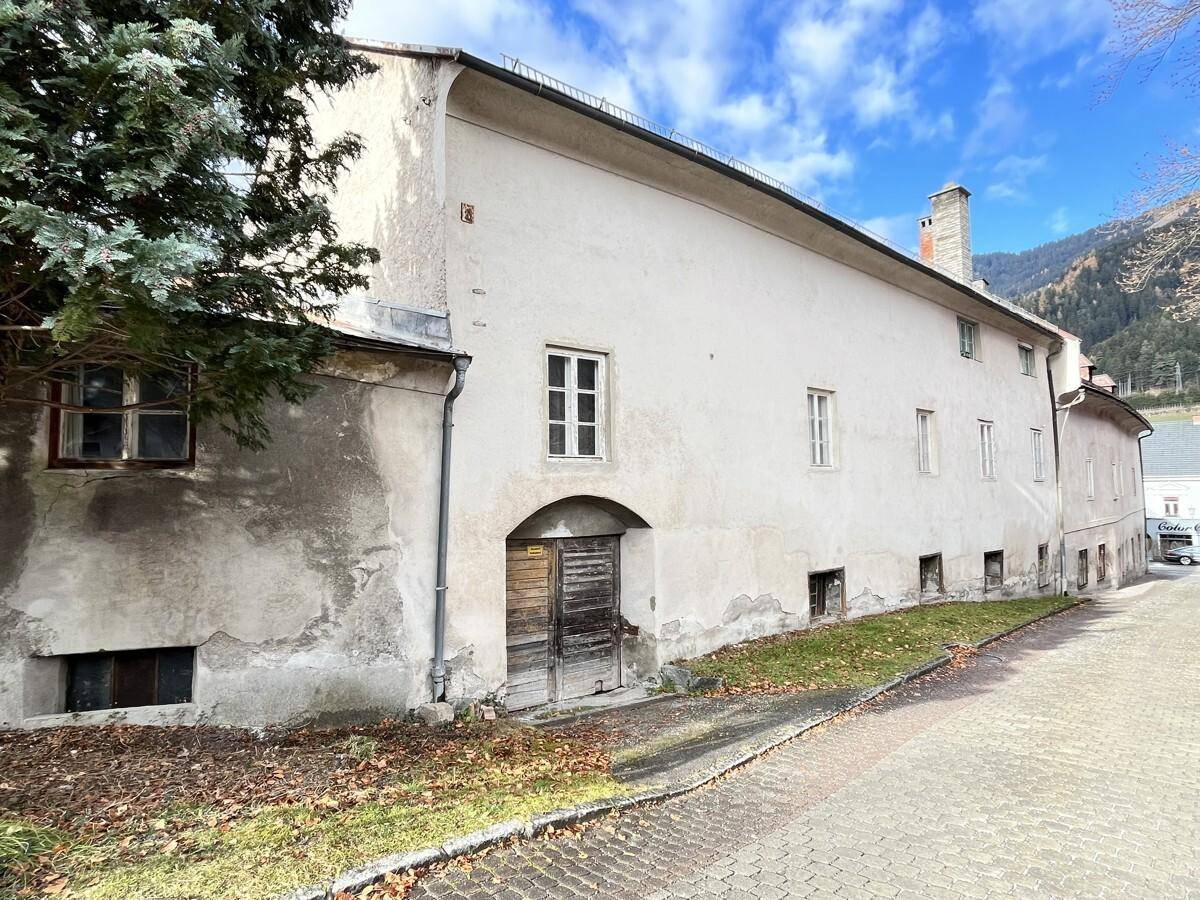 Lange Seitenfassade eines alten Gebäudes mit kleinen Fenstern und einem großen Holztor, umgeben von Grün.