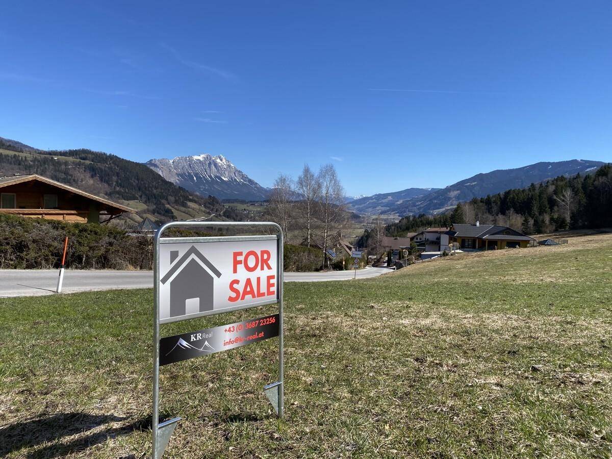 Grundstück mit 'Zu verkaufen'-Schild in einer ländlichen Umgebung mit Bergblick unter blauem Himmel.
