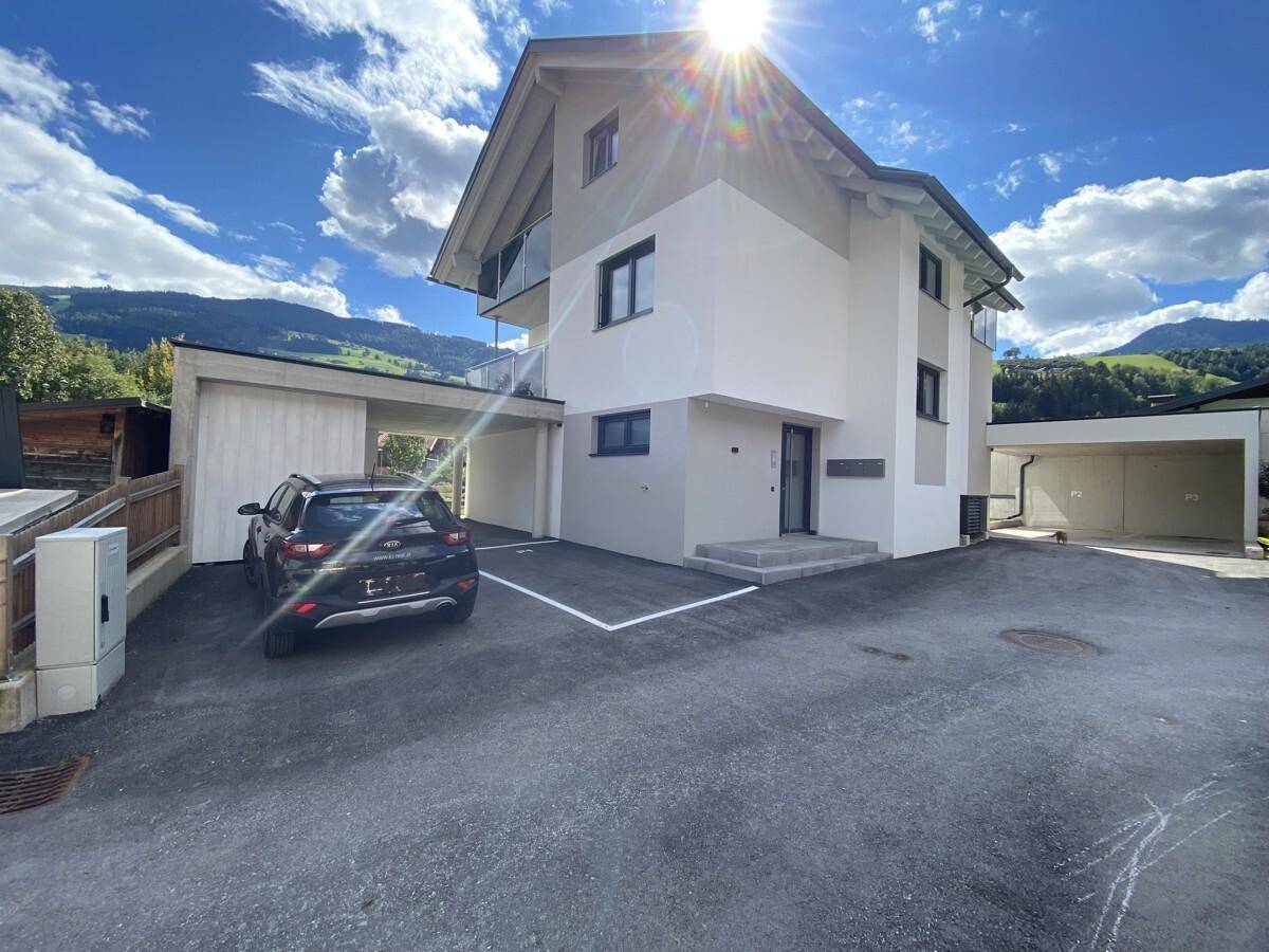 Neubau mit moderner Architektur, Carport und Parkplätzen, umgeben von Berglandschaft.