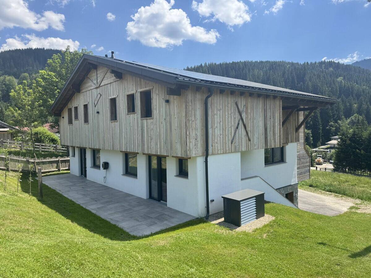 Rückseite des modernen Holzhauses mit Terrasse, Photovoltaikanlage auf dem Dach und Bergblick.