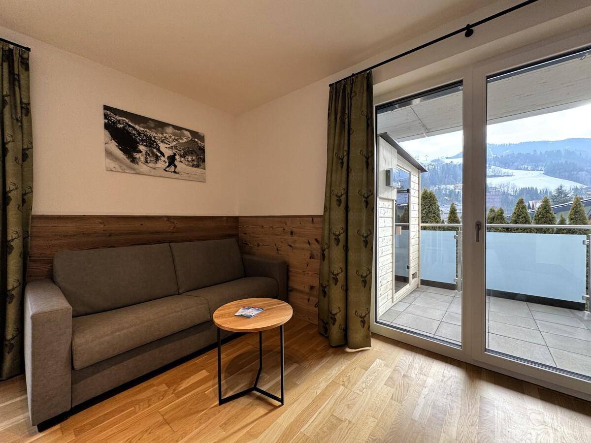 Wohnzimmer mit Sofa, Holzwand und Zugang zum Balkon mit Außensauna und Bergblick.