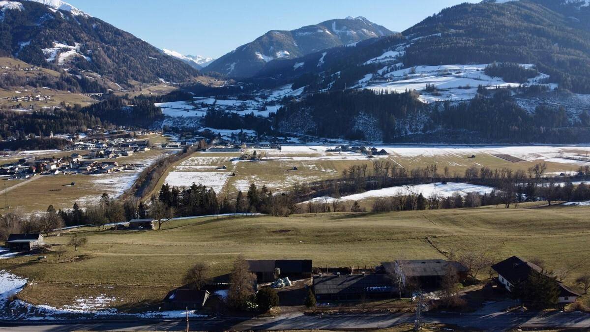 Weitwinkel-Luftaufnahme eines Tals mit Dorf, Feldern, Fluss und schneebedeckten Bergen.