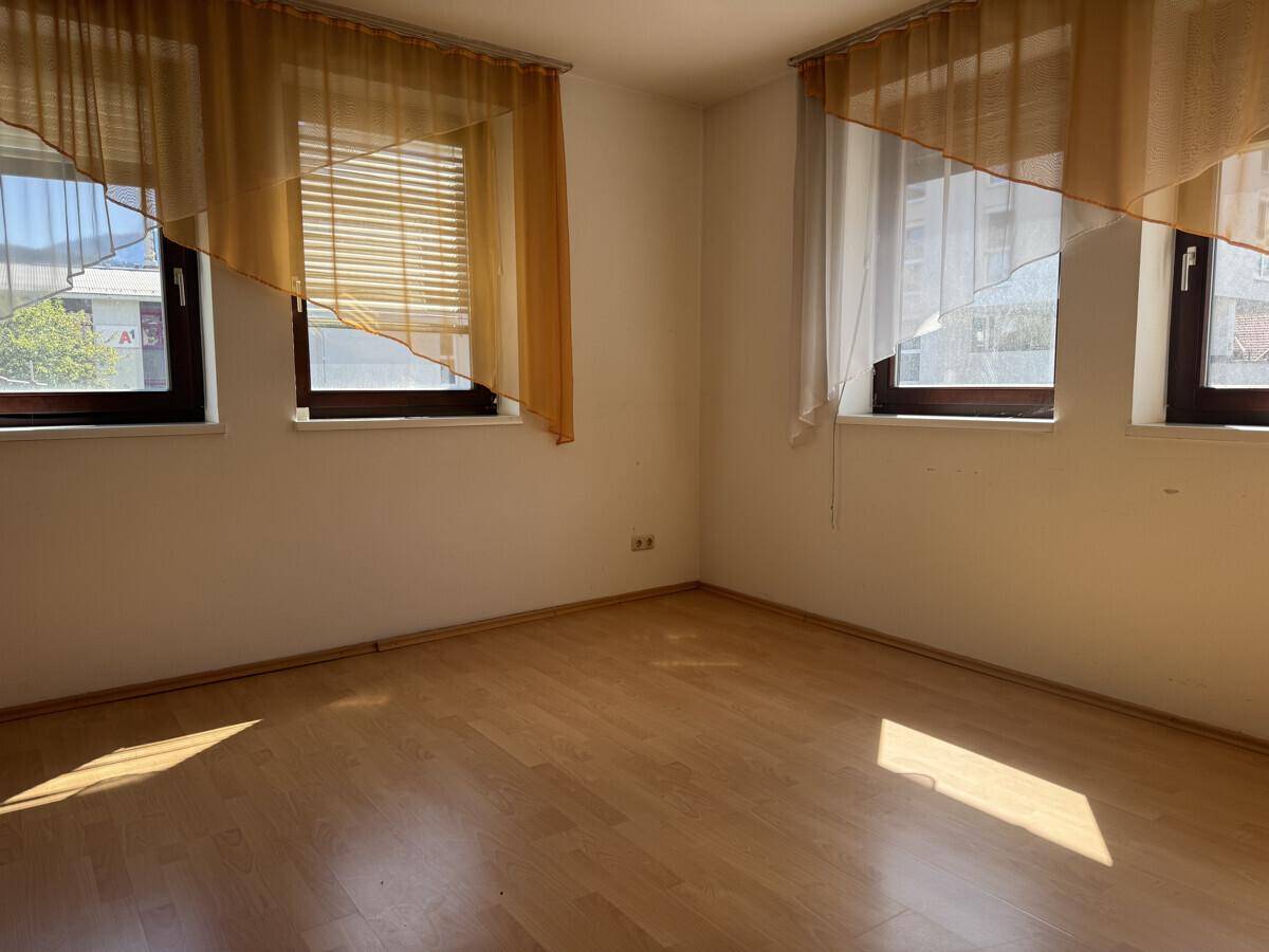 Leeres Schlafzimmer mit Laminatboden, drei Fenstern und gelben Vorhängen.
