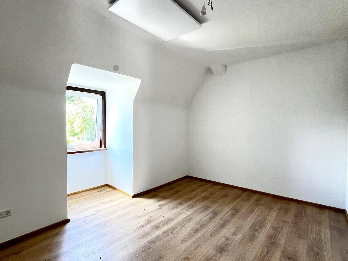 Helles Zimmer mit Laminatboden, weiß gestrichenen Wänden und einem Fenster mit Blick ins Grüne.