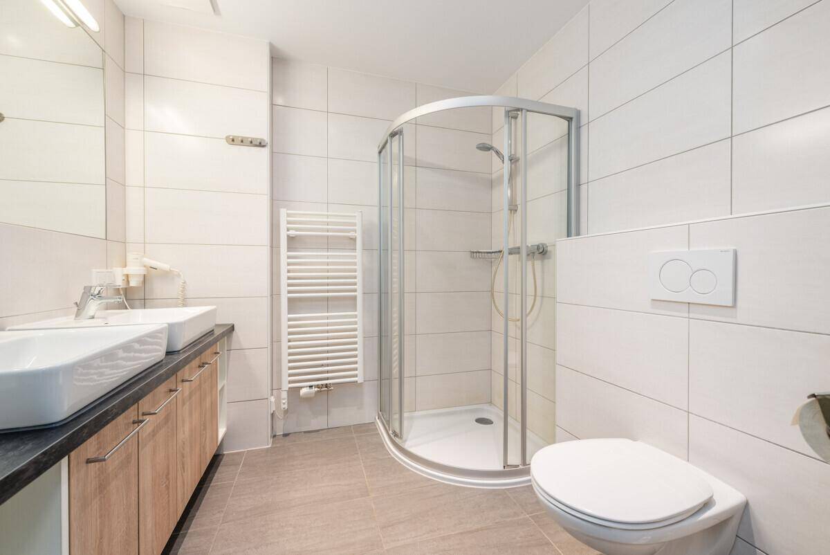 Helles Badezimmer mit Toilette, Doppelwaschbecken, Duschkabine und Handtuchheizkörper.
