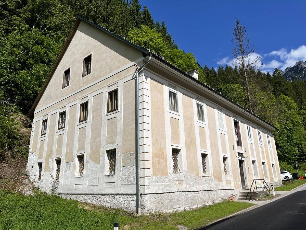 Historisches Gebäude mit Putzfassade und traditionellen Fenstern vor bewaldetem Hang.