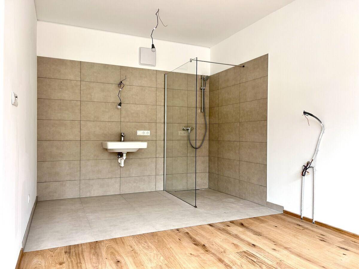 Geräumiges Badezimmer mit begehbarer Dusche, Fliesen und Waschbecken, bereit für die Endmontage.