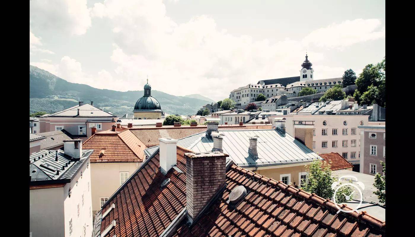 Blick über die Dächer einer historischen Stadt mit Bergen im Hintergrund unter bewölktem Himmel.