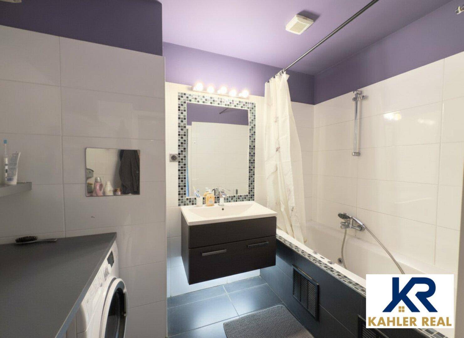 Modernes Badezimmer mit Badewanne, Waschbecken und Waschmaschine, akzentuiert durch lila Decke.
