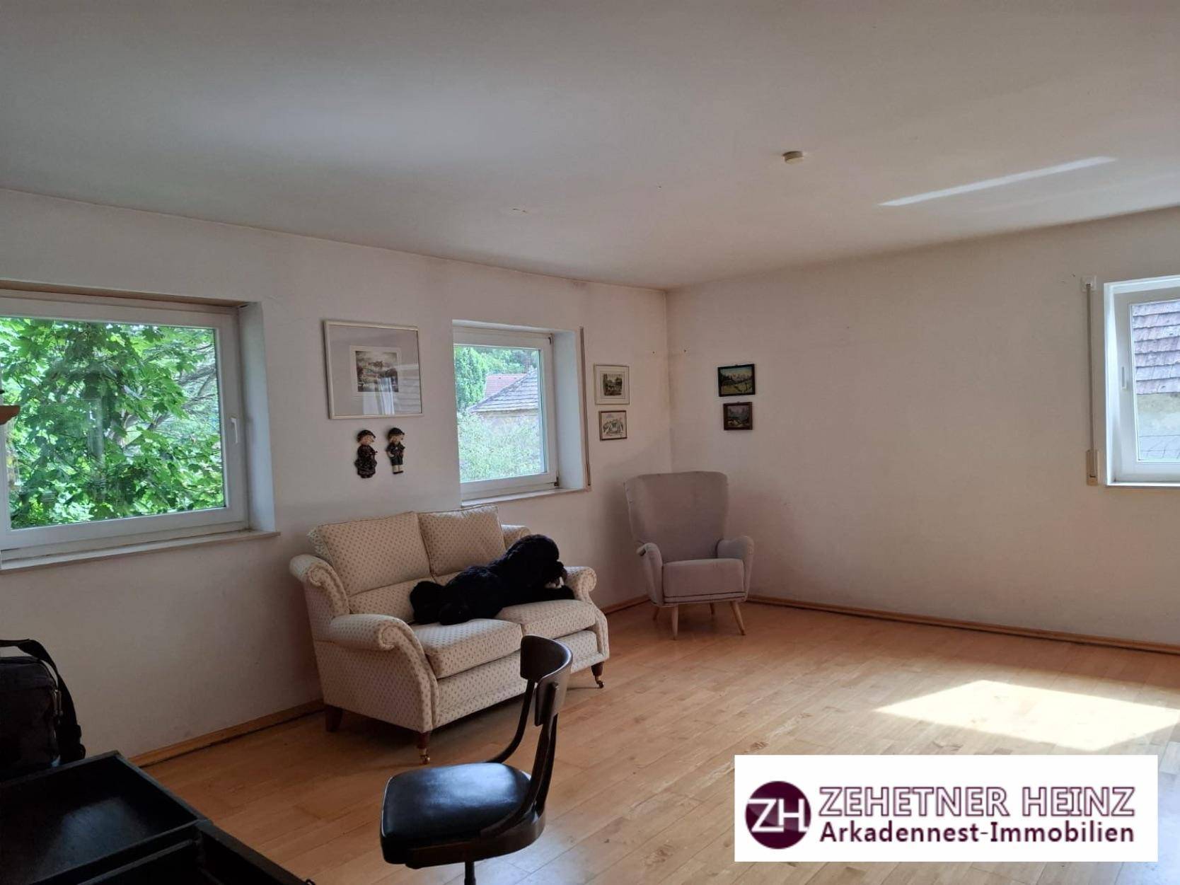 Helles Wohnzimmer mit Holzboden und zwei Fenstern mit Blick ins Grüne.