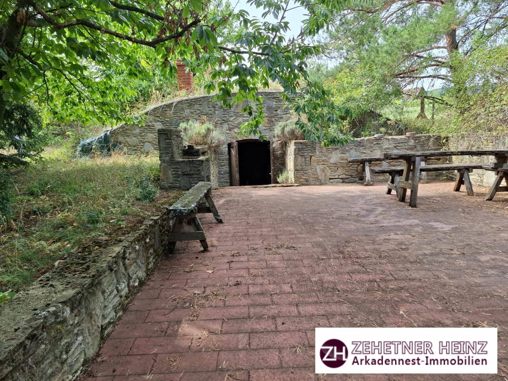 Rustikaler Außenbereich mit gepflasterter Terrasse, Steinmauern und Holzbänken vor einem Erdkeller.