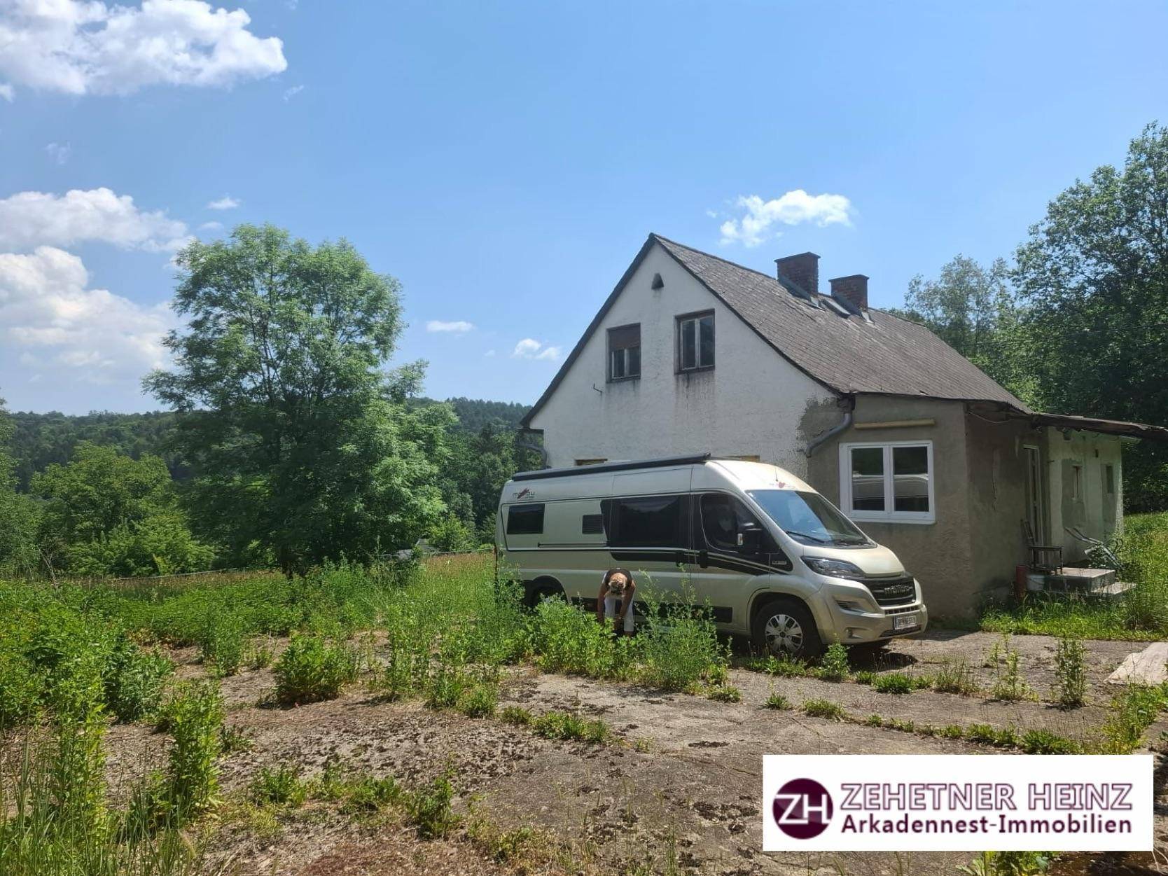 Älteres, einfaches Haus mit Giebeldach und einem geparkten Van, umgeben von überwucherter Vegetation.