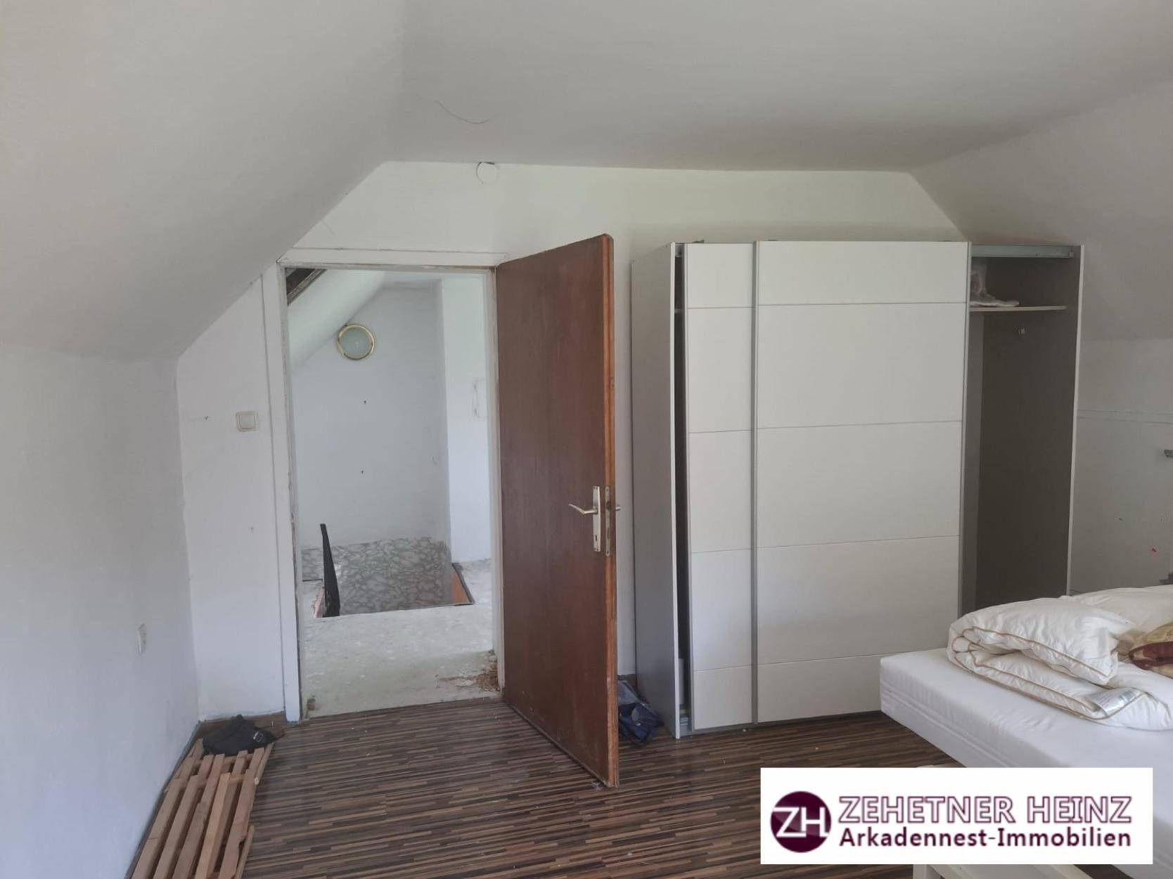 Schlafzimmer mit Bett, weißem Kleiderschrank und offener Tür zu einem weiteren Raum mit Treppe.