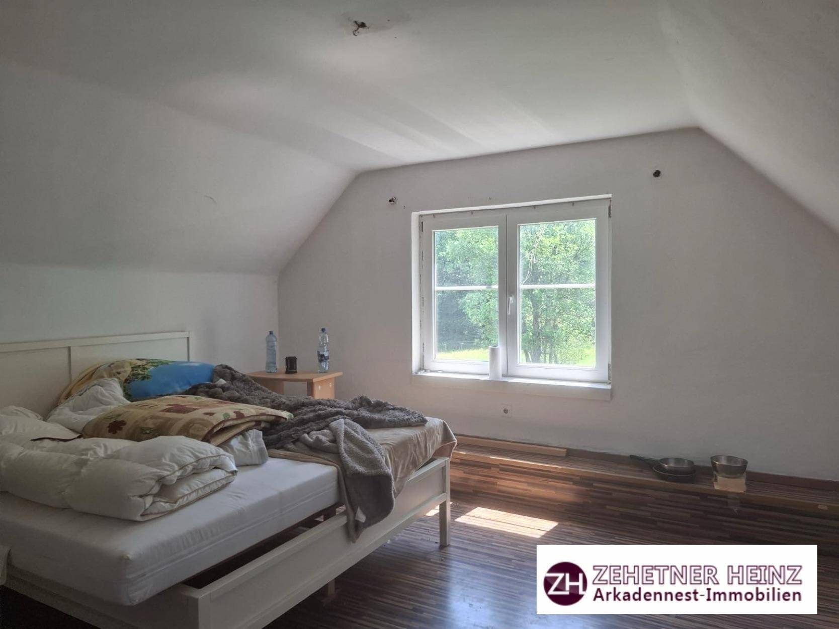 Schlafzimmer mit Bett, Fensterblick auf Bäume und einer Dachschräge, der Boden ist aus Holz.