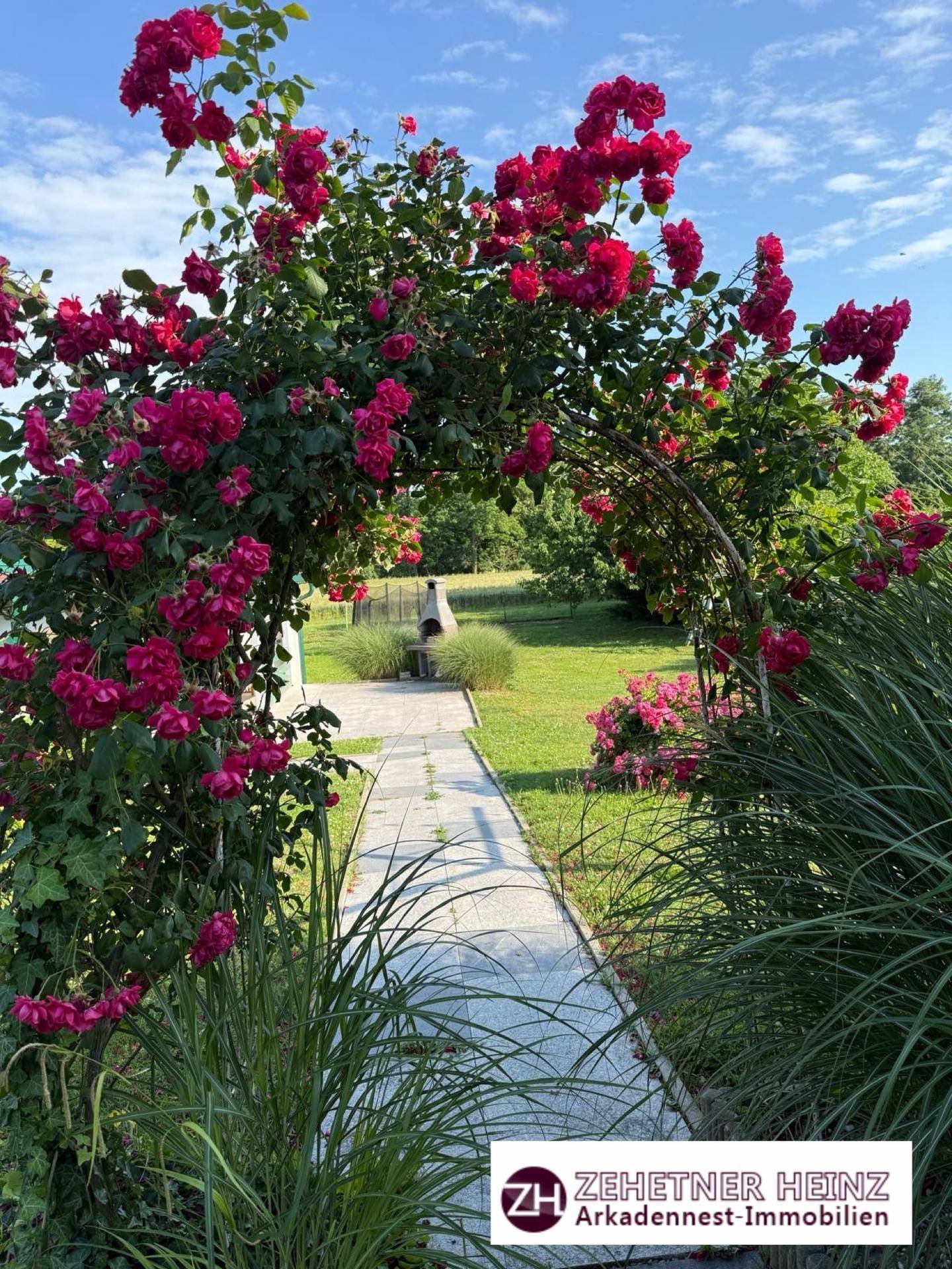 Gepflasterter Gartenweg unter einem Rosenbogen, der zum Grillplatz im Grünen führt.