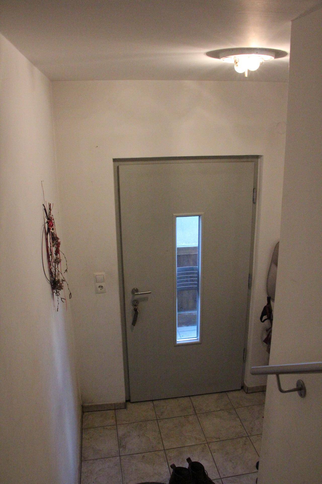 Eingangsbereich mit einer modernen grauen Tür, die ein schmales Fenster besitzt, und Fliesenboden.