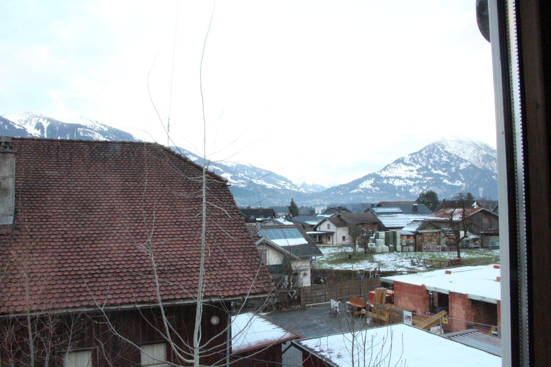 Ausblick aus einem Fenster auf Häuser mit Ziegeldächern und schneebedeckte Berge in der Ferne.