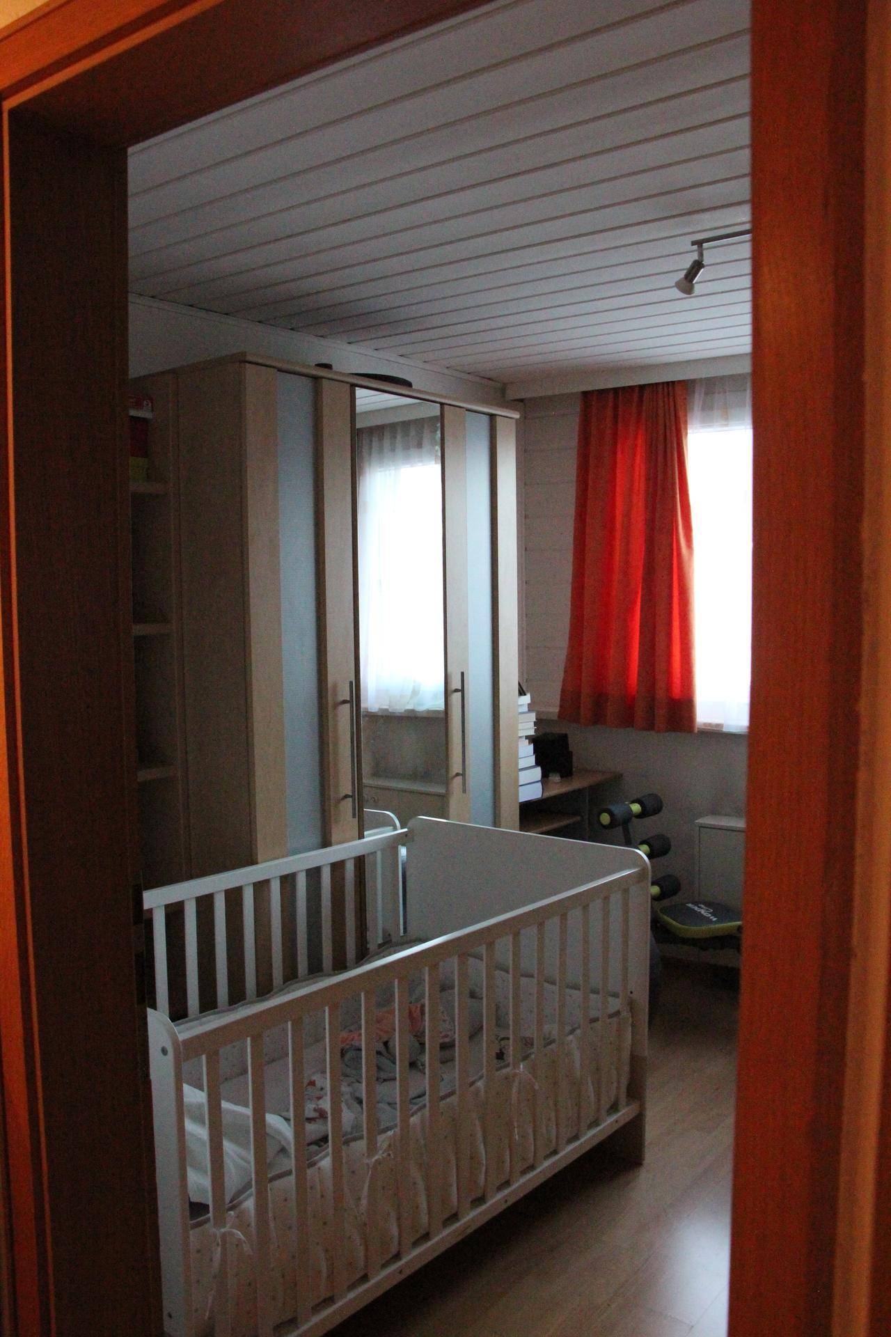 Kinderzimmer mit Babybett, Kleiderschrank und einem Fenster mit roten Vorhängen.