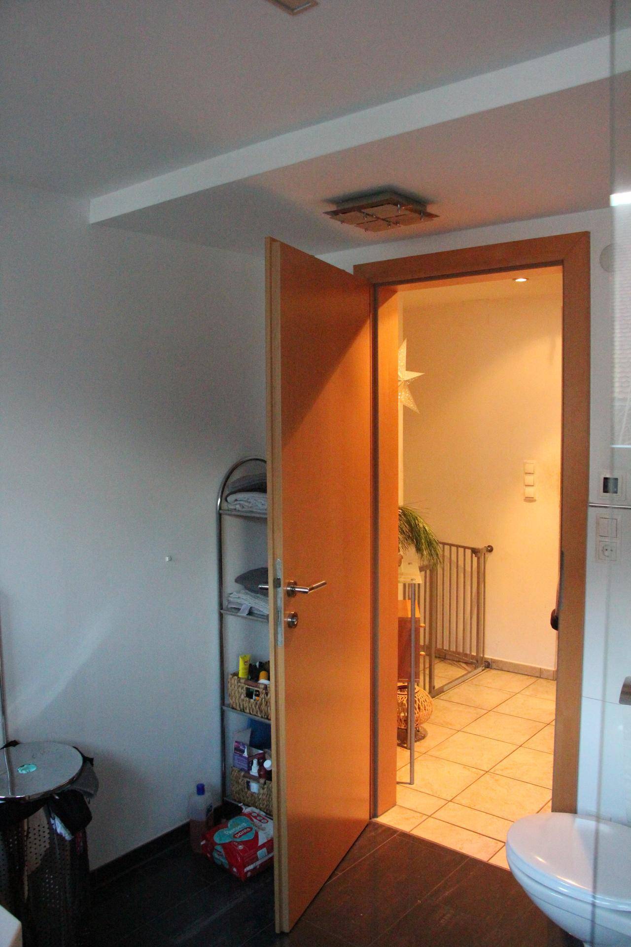 Blick aus dem Badezimmer in den Flur, mit offener Holztür und hellen Fliesenböden.