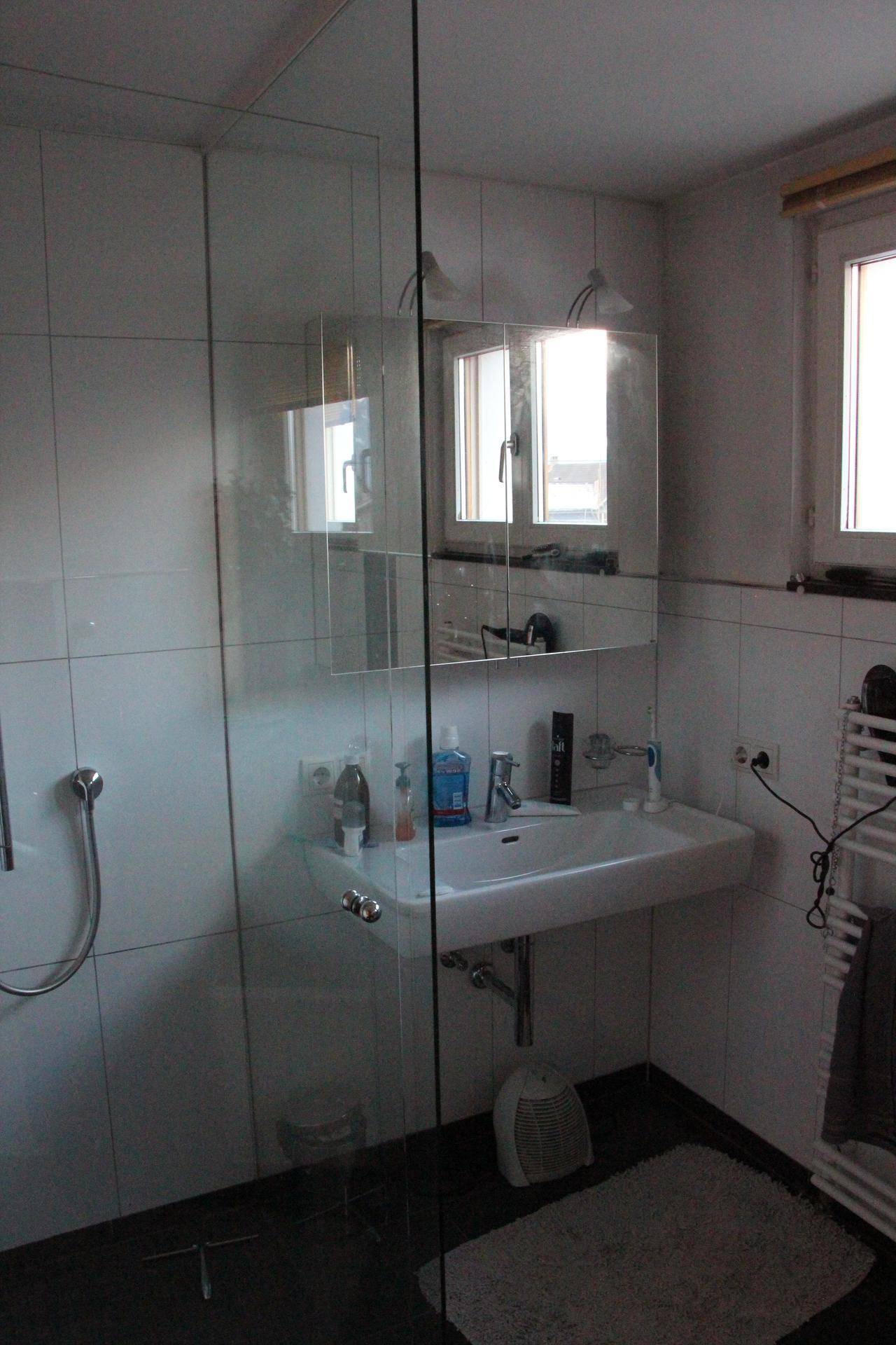Waschbeckenbereich im Badezimmer mit Spiegelschrank, weißen Fliesen und einem Fenster für natürliche Belichtung.
