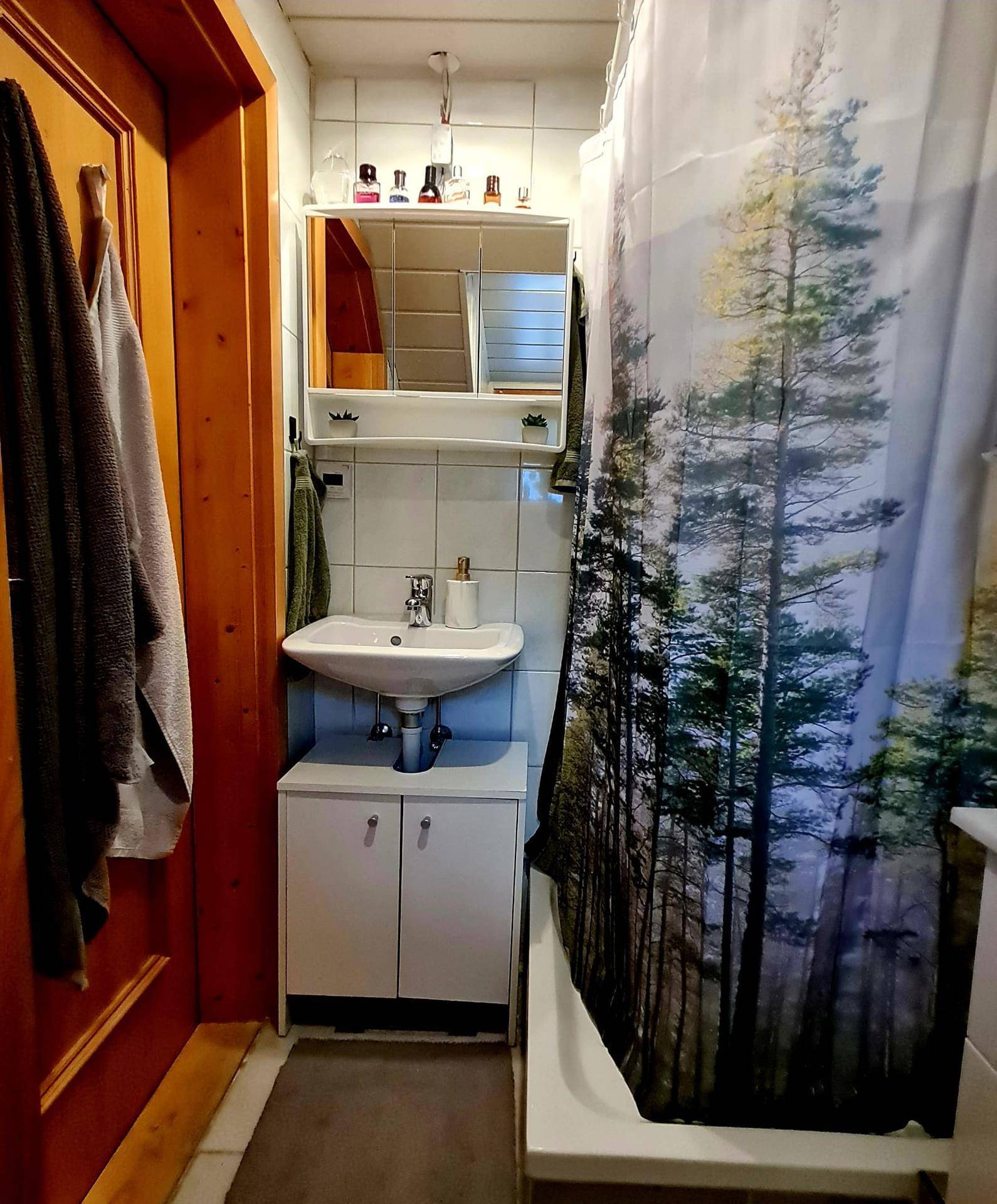 Badezimmer mit Waschbecken, Spiegelschrank und Dusche mit Waldmotiv-Duschvorhang.