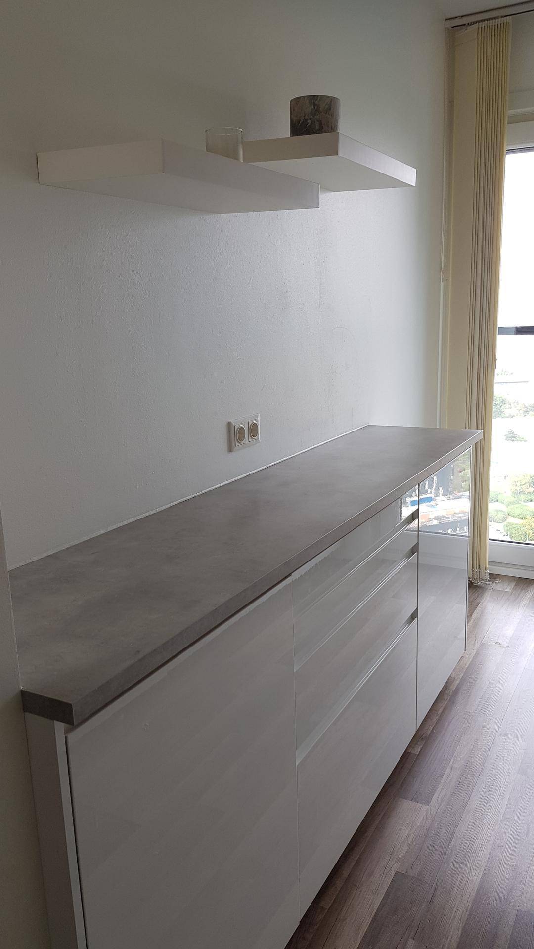 Moderner weißer Schrank mit grauer Arbeitsplatte und schwebenden Regalen neben einem Fenster.