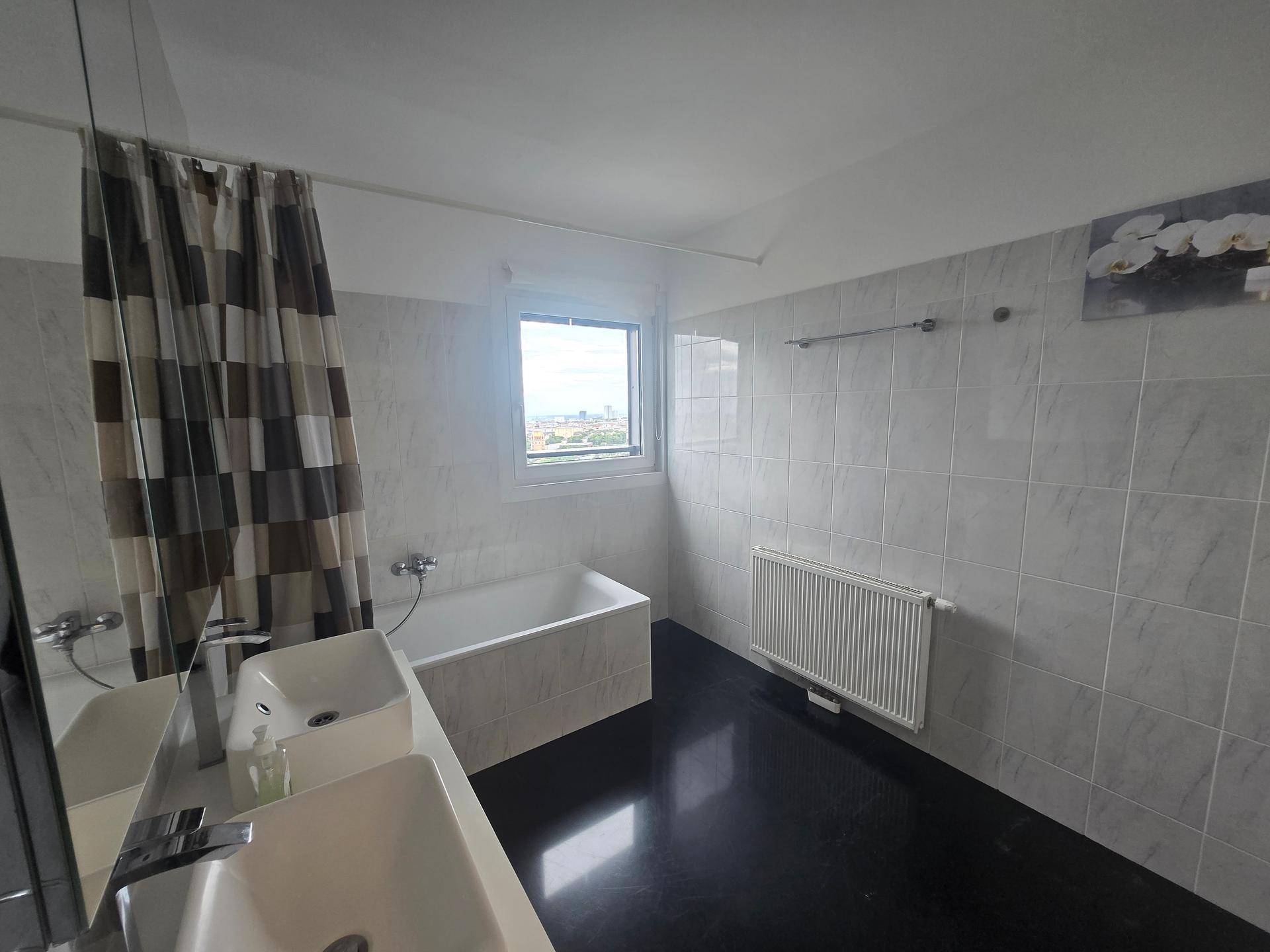 Helles Badezimmer mit Badewanne, Fenster und Heizkörper, umgeben von hellen Fliesen.