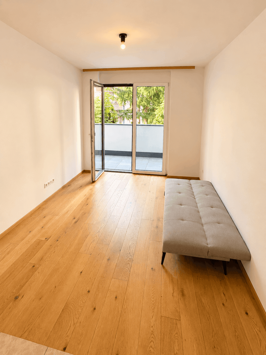 Heller Raum mit Holzboden, Zugang zum Balkon und Blick ins Grüne.