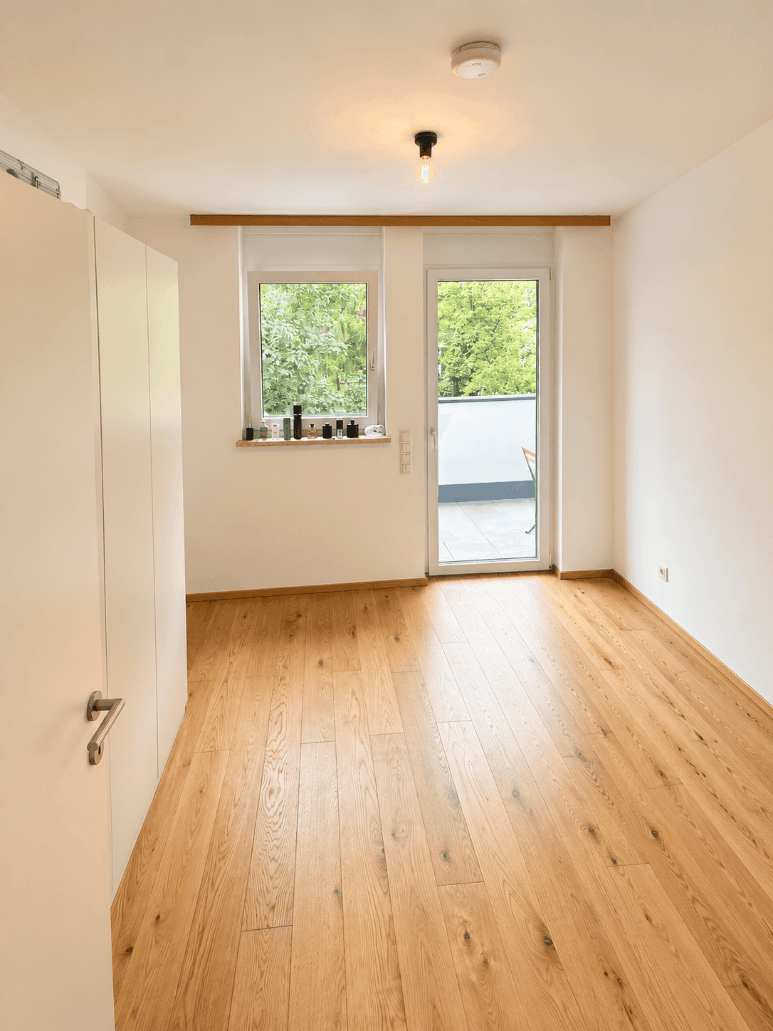 Helles Zimmer mit Holzboden, Fenster und direktem Zugang zum Balkon.