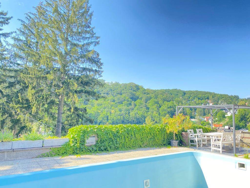 Außenpoolbereich mit gepflasterter Terrasse, Essbereich unter einer Pergola und Blick auf grüne Hügel.