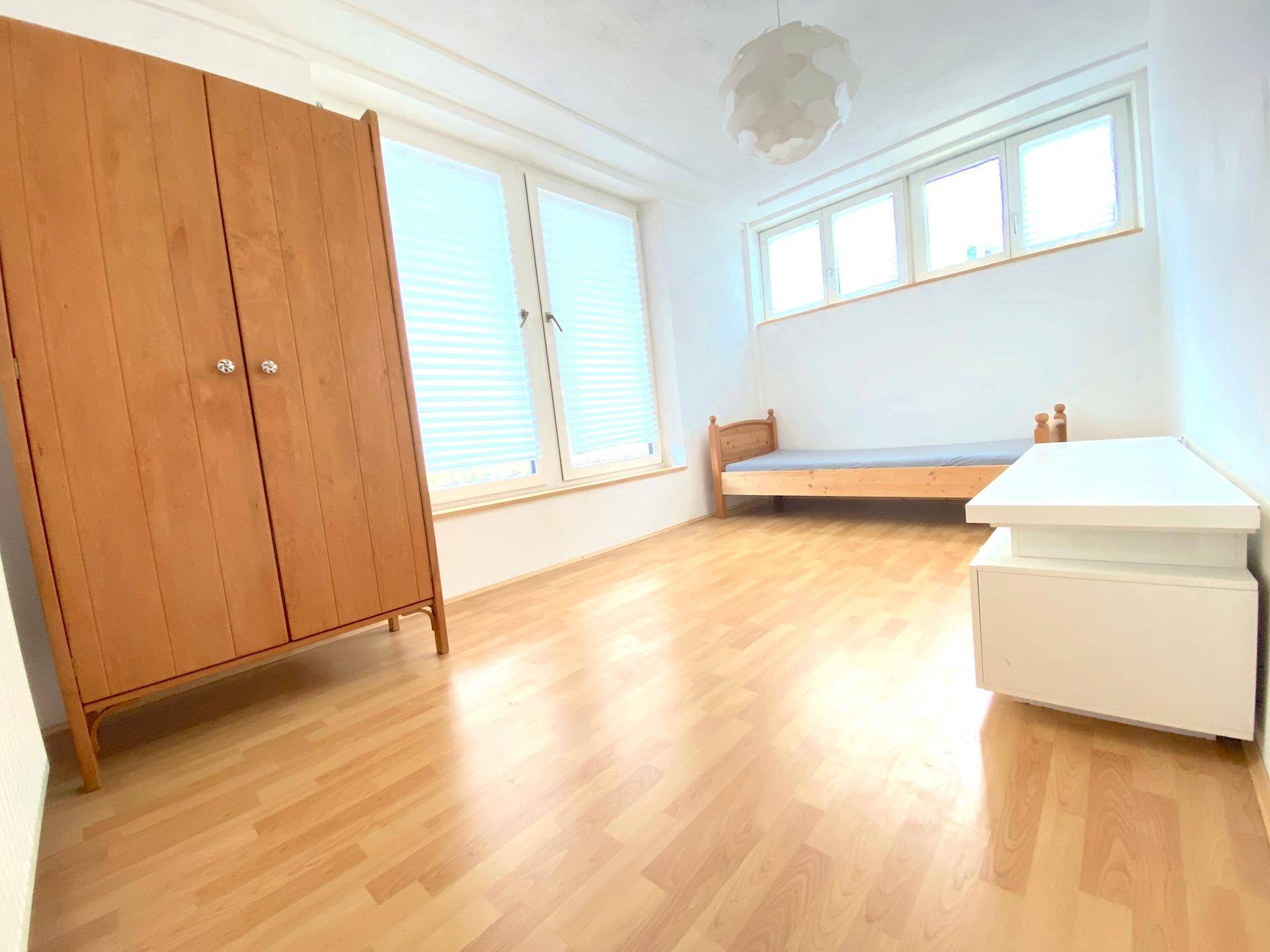 Geräumiges Schlafzimmer mit Holzboden, weiß gestrichenen Wänden und großen Fenstern mit Jalousien.