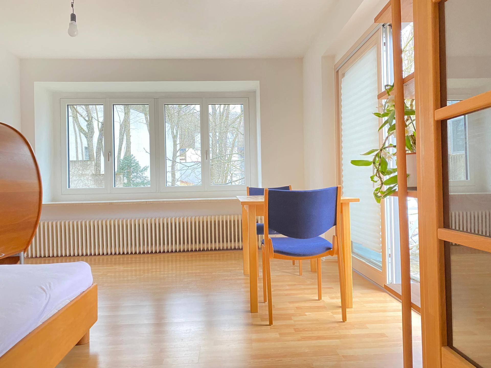 Heller Raum mit Holzboden, großem Fenster und Blick ins Grüne, möbliert mit Tisch und Stühlen.