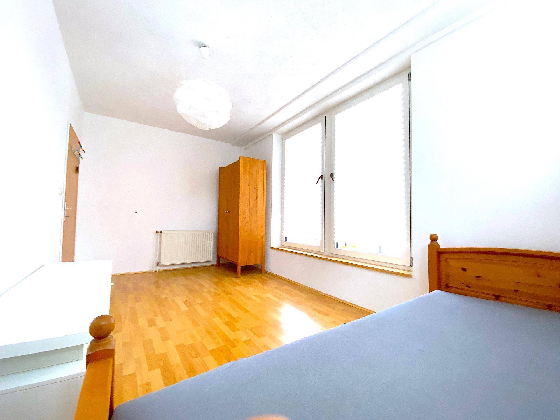 Helles Schlafzimmer mit Holzboden, weiß gestrichenen Wänden und einem Heizkörper unter dem Fenster.